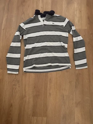Randig långärmad skjorta Lacoste - Säljer en klassisk långärmad skjorta från Lacoste med breda grå och vita ränder. Skjortan har krage, knappar vid halsen och den ikoniska krokodilloggan på bröstet. Perfekt för en avslappnad men ändå stilren look.
