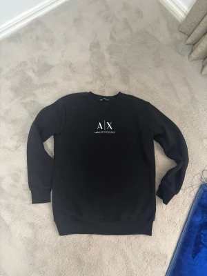 Svart sweatshirt från Armani Exchange - Stilren svart sweatshirt från Armani Exchange med vitt tryck på bröstet. Tröjan har rund halsringning, långa ärmar och ribbade muddar vid ärmslut och nederkant. Perfekt för en clean och modern look.