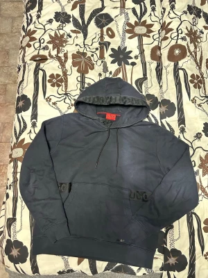 Svart hoodie från HUGO med logga - Säljer en svart hoodie från HUGO med stor huva och dragsnören. HUGO-loggan syns på huvkanten och på fickan framtill. Hoodien har en klassisk känguruficka och är gjord i mjukt bomullsmaterial. Perfekt för en avslappnad och stilren look.