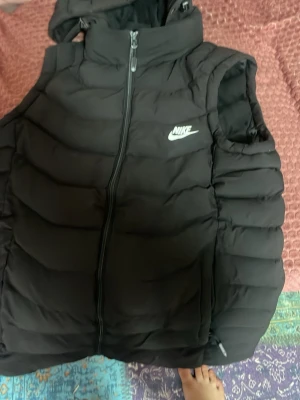 Svart dunväst från Nike - Snygg svart dunväst från Nike med dragkedja framtill och hög krage. Västen har ett vitt Nike-logotyptryck på bröstet och quiltad design för extra värme. Perfekt för lager-på-lager och streetwear-stil.