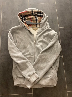 Grå hoodie med Burberry-mönstrad huva - Snygg grå hoodie från Burberry med dragkedja framtill och klassiskt rutigt foder i huvan. Hoodien har två fickor, ribbade muddar och en diskret broderad Burberry-logga på bröstet. Perfekt för en avslappnad men stilren look.
