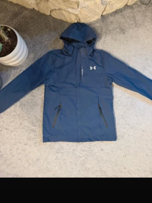 Blå vindjacka från Under Armour - Snygg blå vindjacka från Under Armour med huva och dragkedja framtill. Jackan har två snedställda fickor med dragkedja och ett diskret logotryck på bröstet. Tillverkad i lätt och tåligt material, perfekt för blåsiga dagar.