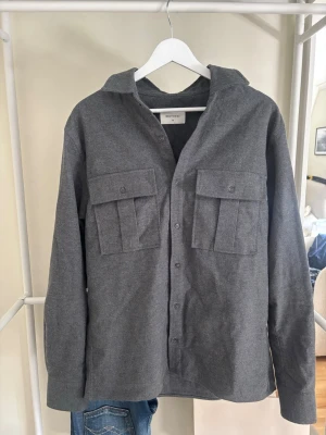 Mörkgrå overshirt med fickor - Stilren mörkgrå overshirt från Brothers med två stora bröstfickor med lock och knappar. Skjortan har klassisk krage, knappstängning framtill och är tillverkad i ett mjukt tyg. Perfekt lager på lager-plagg.
