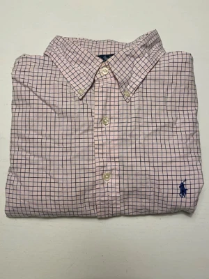 Rutig skjorta från Polo Ralph Lauren - Klassisk rutig skjorta från Polo Ralph Lauren i ljusrosa och blått. Skjortan har knappar framtill, button-down krage och den ikoniska blå broderade loggan på bröstet. Perfekt till jeans eller chinos för en clean och stilren look.