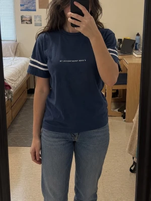 Marinblå t-shirt med vita ränder - Snygg marinblå t-shirt med vita ränder på ärmarna och texten 'BY ASSORTMENT MAD'S' på bröstet. Klassisk passform och rund halsringning. Perfekt till jeans eller shorts för en avslappnad look.