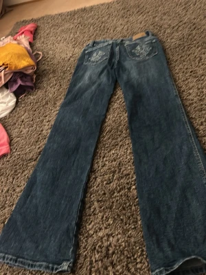 Blå bootcut jeans med broderade fickor - Snygga blå bootcut jeans med broderade detaljer på bakfickorna. Jeansen har klassisk femficksdesign och är tillverkade i denim. Perfekta för dig som gillar en retroinspirerad look med utsvängda ben.