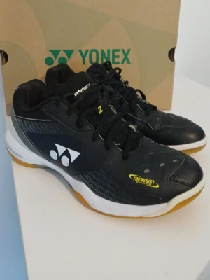 Svarta Yonex inomhusskor - Säljer ett par svarta Yonex inomhusskor med vita och gula detaljer. Skorna har snörning, ventilerande meshpartier och slitstark gummisula. Perfekta för badminton eller andra inomhussporter. Klassisk Yonex-logga på sidan och tåförstärkning. Samma skor som många badminton proffs använder som Viktor Axelsen!