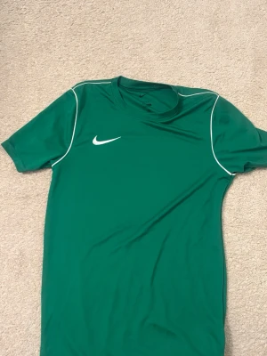 Grön Nike t-shirt  - Snygg grön t-shirt från Nike med vita detaljer och logga på bröstet. T-shirten har korta ärmar och är tillverkad i ett lätt och skönt material, perfekt för sport eller vardag. Klassisk passform och stilren design. 