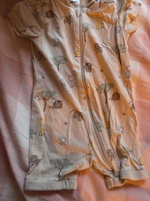 Beige body med djurmönster från Kappahl - Supersöt beige body med dragkedja från Kappahl, storlek 62. Tryck med lejon, giraffer, träd och andra djur i mjuka färger. Korta ärmar och ben, perfekt för små äventyrare. Tillverkad i mjuk bomull för extra komfort.Allt för 100kr