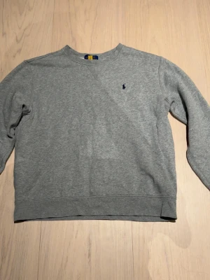 Grå sweatshirt från Polo Ralph Lauren - Klassisk grå sweatshirt från Polo Ralph Lauren med rund halsringning och diskret broderad logga på bröstet. Tröjan har långa ärmar och ribbade muddar vid ärmslut och nederkant. Perfekt för en avslappnad och stilren look.