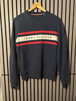 Blå Tommy Hilfiger sweatshirt - Mörkblå sweatshirt från Tommy Hilfiger med klassisk logga och ränder i rött och vitt över bröstet. Använd mycket men bra skick ändå.