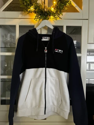Svartvit hoodie från FILA - Säljer en snygg svartvit hoodie från FILA med dragkedja framtill och huva med dragsko. Hoodien har två fickor fram och en broderad FILA-logga på bröstet. Perfekt för en sportig och avslappnad stil.
