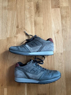 Grå sneakers från Hogan - Snygga grå sneakers från Hogan med mockadetaljer och meshpaneler. Skorna har mörkgrå snören, svart yttersula och en diskret H-logga på sidan. Baksidan har en vinröd detalj som ger extra stilpoäng. Perfekta för dig som gillar klassisk men modern look.