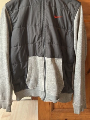Grå hoodie med dragkedja från Nike - Snygg hoodie från Nike i två nyanser av grått med dragkedja framtill och fickor. Framsidan har vadderad panel och ärmarna är i mjukare material. Röd Nike-logga på bröstet och huva med dragsko. Perfekt för en avslappnad streetwear-look.