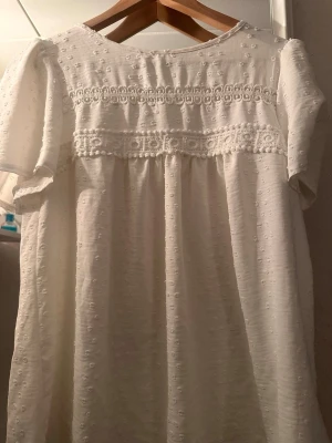 Vit klänning med spetsdetaljer - Söt vit klänning med puffärmar och broderade detaljer. Klänningen har spetsband över bröstet och ett lätt, luftigt tyg som ger en romantisk känsla. Perfekt för varma dagar eller speciella tillfällen.