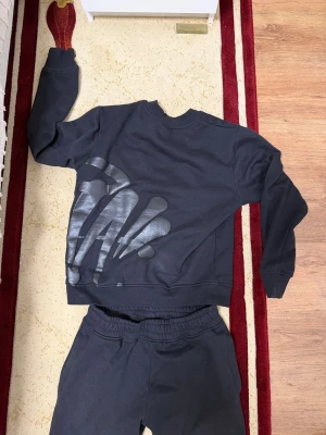 Svart Syna wrld tracksuit - Säljer ett par svarta mjukisbyxor med skön resår i midjan och raka ben. Tillverkade i mjukt bomullsmaterial som passar perfekt för chill dagar. Matcha med en hoodie eller sweatshirt för en avslappnad look.