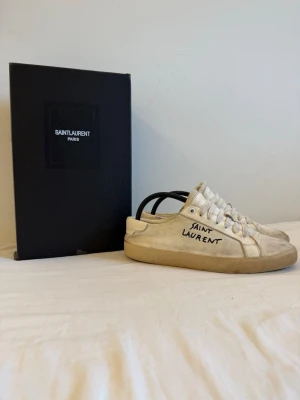  Saint Laurent sneakers - Säljer ett par vita Saint Laurent sneakers med handskriven logga på sidan. Skorna har lågt skaft, vita snören och beige sula. Tillverkade i canvas och läderdetaljer. Klassisk och stilren design med Saint Laurent Paris på plösen. Lite smutsiga men försvinner lätt i tvätten. Pris kan absolut diskuteras 