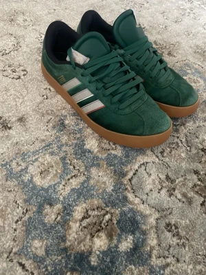 Addidas skor - Snygga gröna sneakers med klassiska vita ränder på sidan och en brun sula. Skorna har lågt skaft och är tillverkade i mockaliknande material med svarta detaljer vid hälen. Perfekta för dig som vill ha en stilren och sportig look.