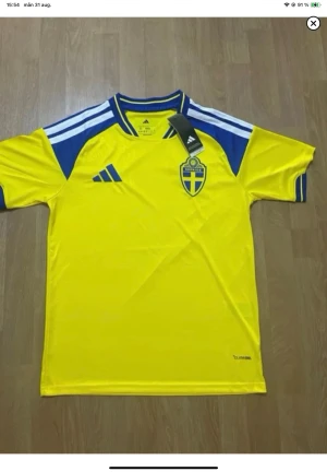 Sverige fotbollströja Adidas gul/blå - Gul fotbollströja från Adidas med blå detaljer och Sveriges landslagsemblem på bröstet. Tröjan har rund hals, blå ränder på axlarna och är tillverkad i ett lätt funktionsmaterial. Perfekt för match eller träning.