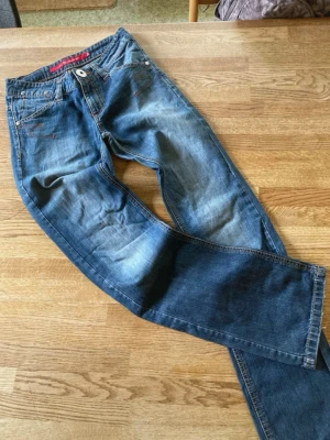 Blå raka jeans med röda detaljer - Säljer ett par blå jeans med raka ben och snygga slitningar. Jeansen har röda sömmar och detaljer vid fickorna samt klassisk femficksdesign. Materialet är denim i bomull och passformen är avslappnad men ändå stilren.