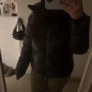 Svart glansig dunjacka från Everest - Snygg svart dunjacka från Everest med glansig finish, huva och två dragkedjeförsedda fickor framtill. Jackan har en puffig look och är perfekt för kalla vinterdagar. Jackan är i strl 40. Den är väldigt varm och har verkligen varit perfekt till vintern. 