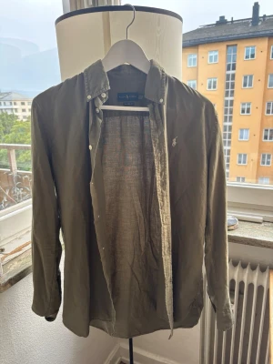 Olivgrön linneskjorta med knappar - Snygg olivgrön skjorta i linne med klassisk krage och knäppning framtill. Skjortan har långa ärmar och en liten broderad logga på bröstet. Perfekt för en avslappnad men stilren look.
