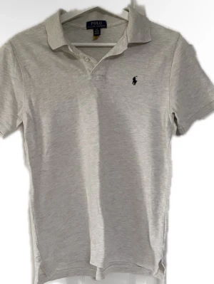 Beige pikétröja från Polo Ralph Lauren - Klassisk beige pikétröja från Polo Ralph Lauren med korta ärmar och krage. Tröjan har knappar vid halsen och den ikoniska broderade loggan på bröstet. Perfekt till jeans eller shorts för en clean look.