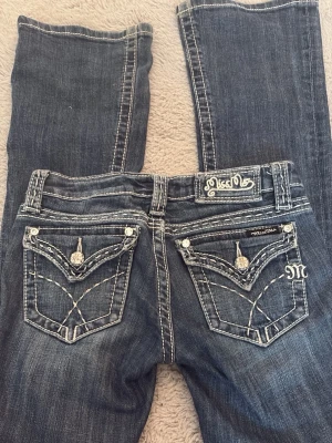 Blå bootcut jeans från Miss Me - Snygga low waist blå bootcut jeans från Miss Me med slitningar och kontrastsömmar. Dragkedjan åker ner men det går att byta