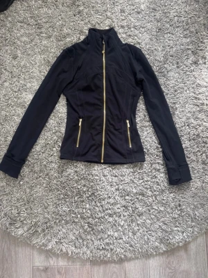lululemon dam  - Snygg svart jacka med figurnära passform och guldiga dragkedjor framtill och på fickorna. Jackan har hög krage och långa ärmar, perfekt för dig som gillar stilrena detaljer och sportig look.