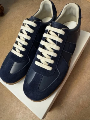  Maison Margiela Gats - Snygga marinblå sneakers från Maison Margiela med ovandel i mocka och läder. Skorna har vita, breda snören och en klassisk låg siluett. Yttersulan är i gummi och insidan är ljus. Perfekta för dig som gillar stilrena och tidlösa sneakers. Aldrig använda helt nya 