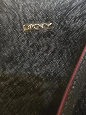 Svart handväska från DKNY - Snygg svart handväska från DKNY i strukturerat läder med guldfärgade detaljer och logotyp framtill. Väskan har en lång axelrem och ett dekorativt DKNY-hänge i guld. Perfekt för dig som vill ha en stilren och trendig accessoar.