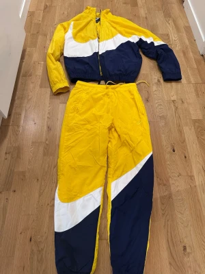 Nike gul och blå tracksuit - Säljer en retro tracksuit från Nike i gult, blått och vitt. Setet består av en jacka med dragkedja och resår i ärmslut samt matchande byxor med dragsko i midjan. Materialet är lätt och passar perfekt till träning eller streetwear.