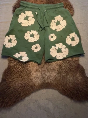 Gröna shorts med vita blommor - Unika gröna shorts med stora vita blommor tryckta över hela plagget. Shortsen har snörning i midjan för justerbar passform och är tillverkade i ett mjukt bomullsmaterial. Perfekta för varma dagar och ger en avslappnad, trendig look.