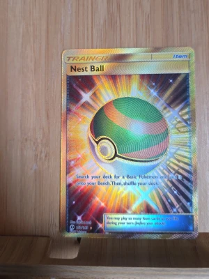Pokémonkort Nest Ball Secret Rare - Säljer ett Pokémonkort, Nest Ball Secret Rare, med guldig ram och holografisk effekt. Kortet har färgstarka detaljer i grönt, orange och guld. Perfekt för samlare eller dig som vill boosta din lek med en snygg tränarkort-item.