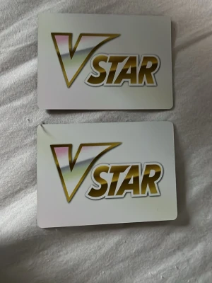 Pokémon VSTAR markörer i plast - Två Pokémon VSTAR markörer i plast med guldfärgad text och vit bakgrund. Ena sidan har VSTAR-loggan i guld och vitt, andra sidan är svart med en Pokéboll-symbol. Perfekt till Pokémon TCG-spel.