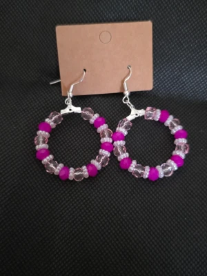 Runda örhängen med rosa pärlor - Säljer ett par snygga örhängen i rund form med genomskinliga och starkt rosa pärlor. Örhängena har metallkrokar i silverfärg och pärlorna är trädda på en cirkelformad stomme. Perfekta för att ge din look en färgklick!
