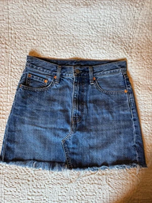 Levis jeanskjol med fransig kant - Säljer en klassisk Levis jeanskjol i kort modell med fransig kant nedtill. Storlek 26 i midjan. Perfekt till sommaren och enkel att styla med olika toppar.