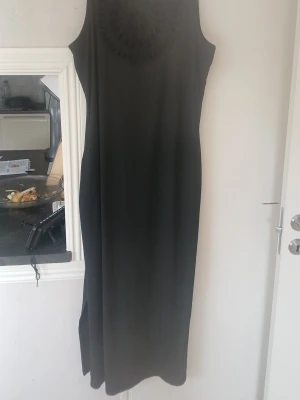 Svart långklänning med spetsdetalj - Stilren svart långklänning med ärmlös design och dekorativ spetsdetalj vid halsringningen. Klänningen har en rak passform och slits i sidan för extra rörelsefrihet. Perfekt för dig som gillar enkel men elegant stil.
