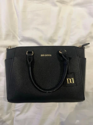 Svart handväska från Don Donna - Snygg svart handväska från Don Donna med guldfärgade detaljer och dubbla handtag. Väskan har en stilren, fyrkantig form och är tillverkad i ett tåligt material med struktur. Perfekt för dig som vill ha en klassisk och trendig accessoar.