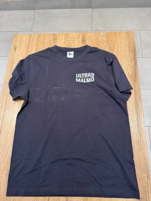 Svart Ultras Malmö t-shirt - Snygg svart t-shirt med trycket 'ULTRAS MALMÖ' på bröstet. Klassisk passform och rund halsringning. Perfekt för dig som vill visa ditt stöd för Malmö. Tillverkad i mjuk bomull för skön känsla hela dagen.