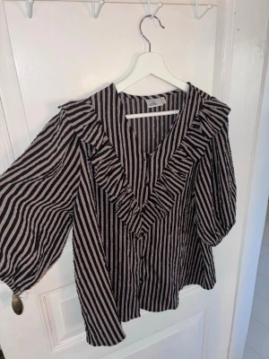 Randig blus med volang från Isolde - Säljer en svart och beige randig blus från Isolde med volangdetaljer längs v-ringningen och knäppning framtill. Blusen har trekvartsärmar och ett luftigt, skrynkligt tyg som ger en avslappnad look.