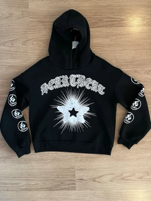 Svart hoodie med tryck och huva - Svart hoodie med stor huva och grafiskt tryck framtill med texten 'HEARTTREAT' och en stjärna i mitten. Ärmarna har vita tryck med symboler längs sidorna. Croppad modell med ribbade muddar. Perfekt för en edgy streetwear-look.