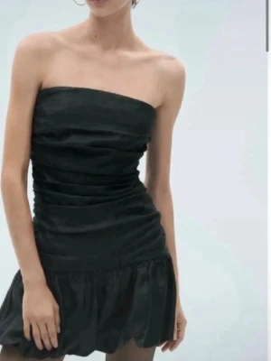 Svart axelbandslös kort klänning - Trendig svart axelbandslös kort klänning med rynkade detaljer och volang nedtill. Klänningen har en figurnära passform och är perfekt för dig som vill ha en stilren och modern look. Tillverkad i ett glansigt material som ger en extra snygg touch.