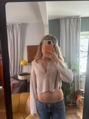 Beige cropped stickad tröja - Beige stickad tröja i croppad modell med långa ärmar och ribbad struktur. Tröjan har en djup v-ringning med metall knappar och avslappnad passform. Perfekt att styla med jeans för en chill look.