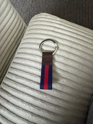 Nyckelring med blått och rött band - Snygg nyckelring med metallring, brunt läderfäste och ett band i marinblått med en röd rand i mitten. Perfekt accessoar för att hålla koll på nycklarna och ge en stilren touch till din vardag.