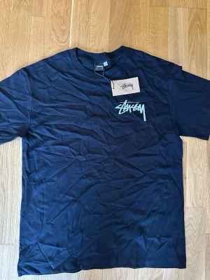 Svart Stüssy t-shirt med tärningsmotiv - Säljer en svart t-shirt från Stüssy med deras ikoniska logga på bröstet och ett stort tryck med två tärningar på ryggen. T-shirten har korta ärmar och är gjord i mjuk bomull. Perfekt för dig som gillar streetwear och vill sticka ut.