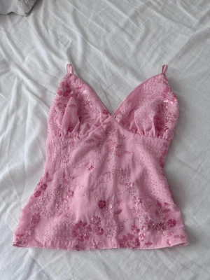 Rosa topp med paljetter och broderi - Supersöt rosa topp med tunna axelband, djup urringning och vackra paljetter samt broderade blommor över hela plagget. Figurnära passform och lätt glansigt material som ger en lyxig känsla. Perfekt för att lyfta vilken outfit som helst.