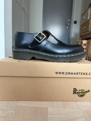Svarta Dr. Martens T-bar loafers - Snygga svarta loafers från Dr. Martens med klassisk T-bar design och spänne i metall. Skorna har robust sula med gul söm och är tillverkade i slätt läder. Perfekta för dig som vill ha en edgy och tidlös stil.