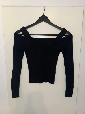 Svart off shoulder topp med långa ärmar - Trendig svart stickad topp med off shoulder-design och breda axelband. Toppen har figurnära passform och långa ärmar, perfekt för en stilren look. Materialet är mjukt och stretchigt för extra komfort.