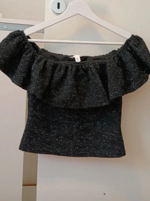 Svart glittrig offshoulder topp - Supersnygg svart offshoulder topp med glittrigt tyg och stor volang runt axlarna från H&M, endast använd 1 gång. Skriv om du har någon fråga 🤗